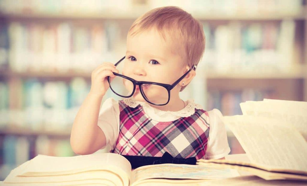 An infant sits at a table in a library with books in front of her. She is wearing a plaid top and has a pair of oversized black glasses that she is holding in front of her face.
